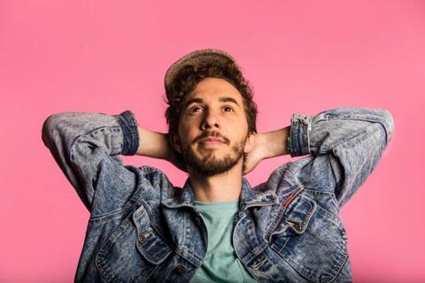 Un hombre con chaqueta de mezclilla y gorra posando frente a un fondo rosa.