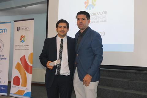 Dos hombres posan juntos en un evento de graduación, uno de ellos sostiene un premio.