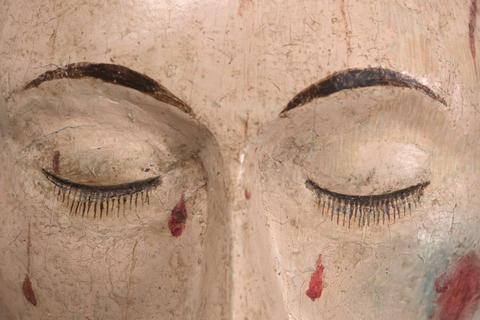 La imagen muestra un rostro de madera con los ojos cerrados y marcas de color rojo.