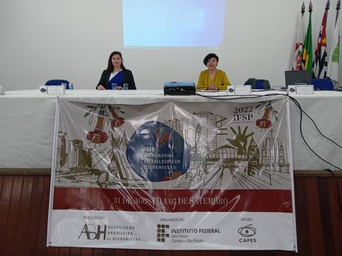 Imagen de dos mujeres sentadas en una mesa frente a un banner de un congreso.