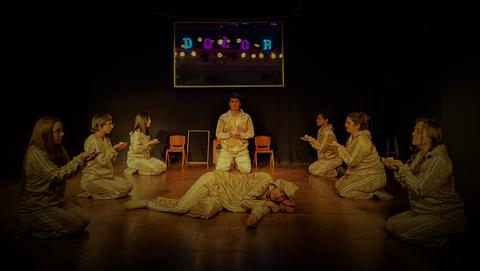 Un grupo de actores en un escenario representan una escena teatral con un personaje acostado en el suelo.