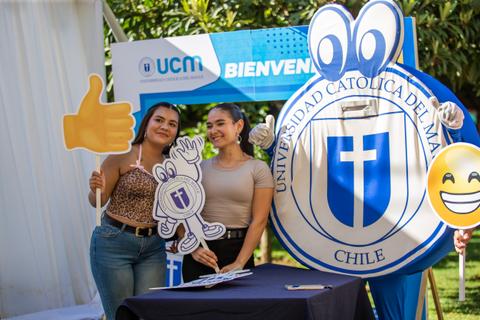 Dos jóvenes posan sonrientes con carteles y un logo de la universidad en un evento de bienvenida.