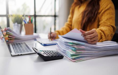 Una persona revisando documentos en un escritorio desordenado con una laptop y una calculadora.