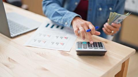 Una persona usando una calculadora mientras sostiene dinero y revisa documentos.