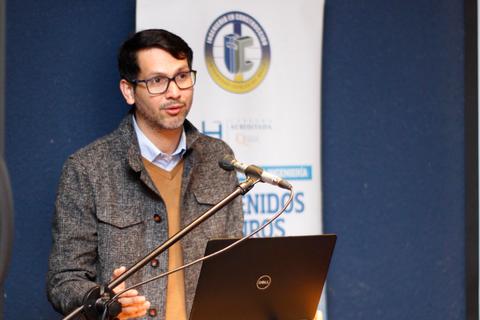 Un hombre está dando una charla en un evento, de pie frente a un micrófono y una computadora portátil.
