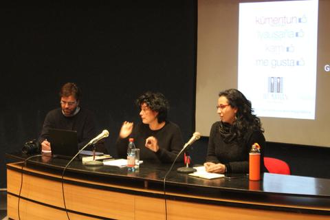 Una charla donde tres personas discuten en un escenario con micrófonos y una presentación detrás.