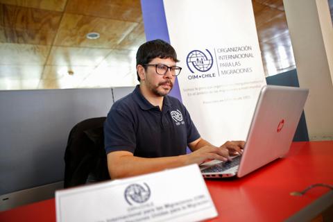 Un hombre está trabajando en su computadora portátil en un entorno de oficina, al lado de un cartel de la OIM-Chile.