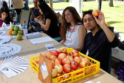 Un grupo de jóvenes se encuentra en una mesa, donde uno de ellos sostiene una manzana y hay una cesta llena de manzanas.