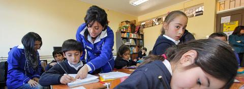 Un aula con niños y una maestra que les asiste en sus tareas escolares.