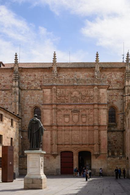La imagen muestra la fachada de un edificio histórico con una estatua en primer plano y personas caminando.