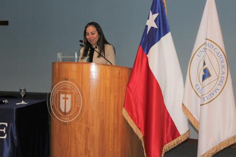 Una mujer se encuentra dando una charla en un podio frente a la bandera de Chile y un estandarte de la universidad.