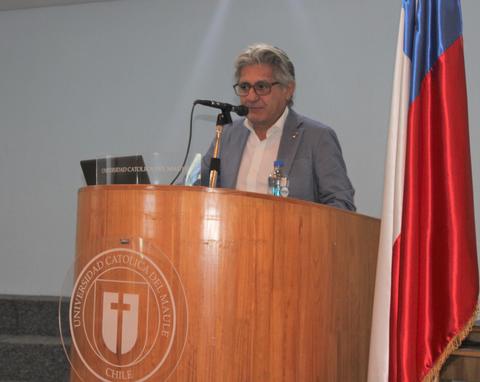 Un hombre presenta en un podio de una universidad con la bandera chilena al fondo.