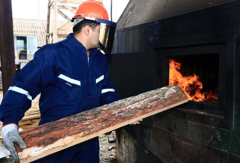 Un hombre vestido con un overol azul y casco naranja está introduciendo un tronco en un horno industrial donde hay fuego.