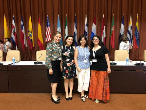 Cuatro mujeres posando en un evento, con banderas de varios países de fondo.