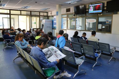 Una sala de espera con varias personas sentadas, algunas mirando televisión y otras leyendo un periódico.