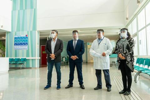 Cuatro personas, vestidas formalmente y usando protectores faciales, están de pie en un entorno interior que parece ser un hospital.