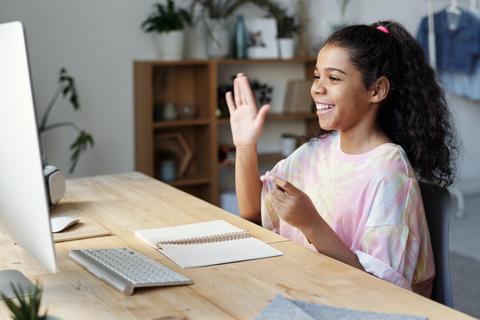 Una niña sonriente saluda frente a un ordenador en un ambiente acogedor.