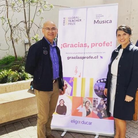 Dos personas posan junto a un cartel que agradece a los profesores y promueve un premio internacional de enseñanza.