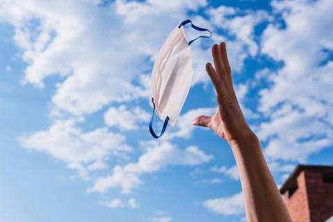 Una mano lanza una mascarilla blanca al cielo despejado.