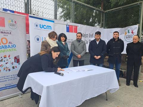 Un grupo de personas asisten a un evento donde una mujer está firmando un documento en una mesa.