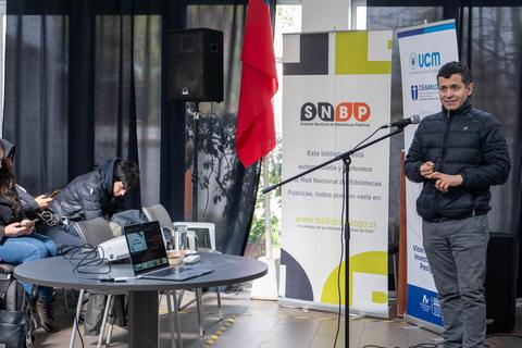 Un orador presenta en un evento rodeado de público y material informativo.