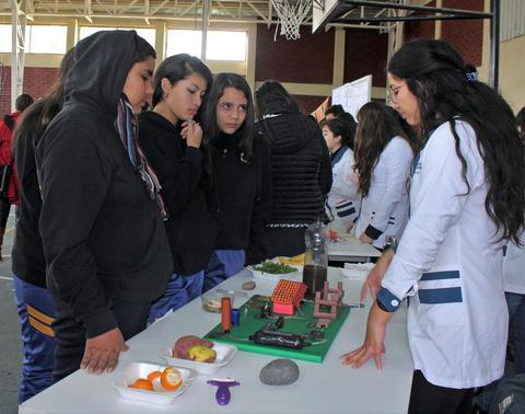 Un grupo de estudiantes observa una presentación de proyectos en una mesa en un ambiente escolar.