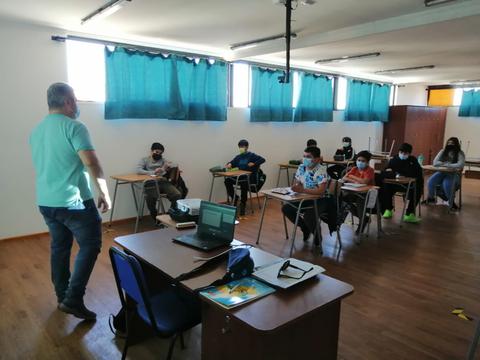 Una clase en un aula con estudiantes prestando atención al profesor que está de pie.