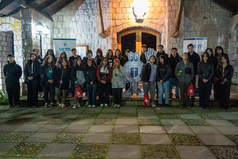 Un grupo de personas posando frente a un edificio iluminado, con banderas y una mascota en el fondo.