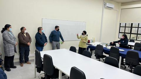 Un grupo de personas se encuentra en una sala de reuniones, escuchando a una mujer que está hablando frente a un pizarrón.
