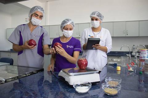 Tres personas en un laboratorio de alimentos analizan manzanas en una balanza.