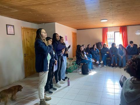 Un grupo de personas en una sala, algunas de pie y otras sentadas, escuchando una presentación.