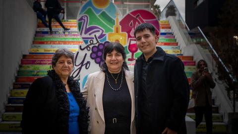 Tres personas posan frente a una colorida escalera decorada con imágenes y frases relacionadas con el vino y la cultura local.