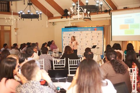 Una conferencia con una presentadora en un salón lleno de asistentes sentados frente a ella.