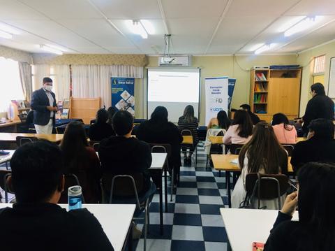 Un grupo de estudiantes asiste a una presentación en un aula con un expositor frente a una pantalla.