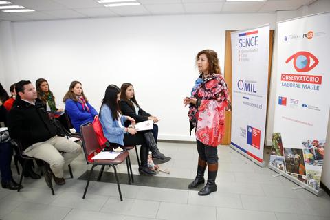 Una mujer presenta ante un grupo de personas en un aula con carteles de diferentes organizaciones en las paredes.