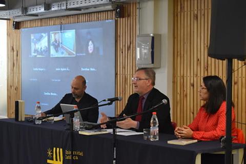 Una mesa de discusión con varios ponentes frente a una pantalla con participantes virtuales.