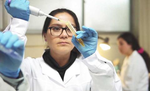 Una mujer con guantes azules y bata de laboratorio mide un líquido con una pipeta en un entorno de laboratorio.