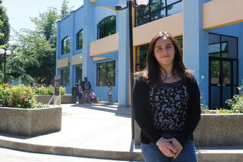 Una joven sonríe frente a un edificio de color azul con jardines y personas al fondo.