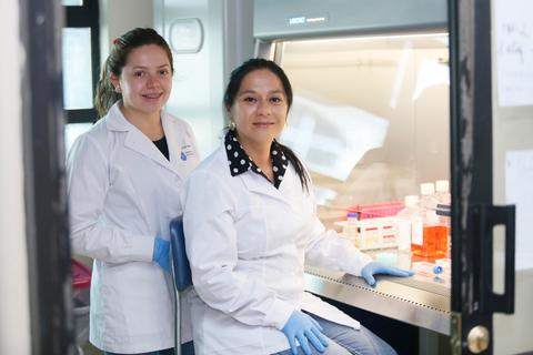Dos mujeres científicas sonríen mientras están en un laboratorio con equipos de análisis y frascos de muestra.