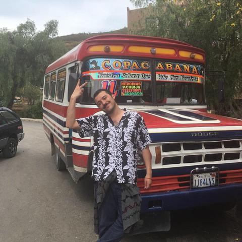 Un hombre sonriente con una camisa de flores posando frente a un autobús colorido con decoraciones llamativas.