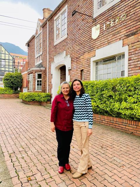 Dos mujeres posan sonrientes frente a un edificio de ladrillo en un día nublado.