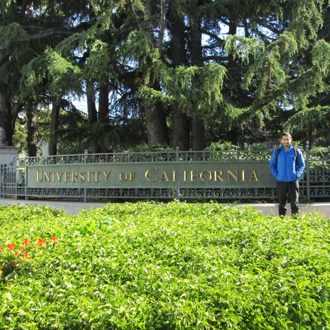 Una persona se encuentra de pie junto a un cartel que dice 'University of California', rodeado de vegetación.