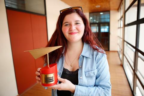 Una joven con cabello rojizo sonríe mientras sostiene un trofeo en forma de avión en un pasillo interior.