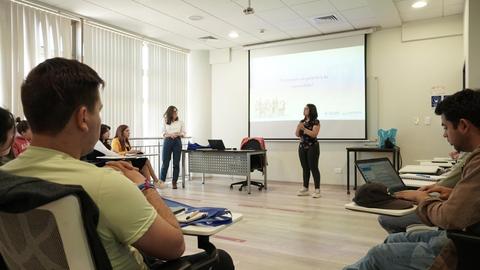 Un grupo de personas en una sala de clases escucha dos presentadoras en un proyecto educativo.
