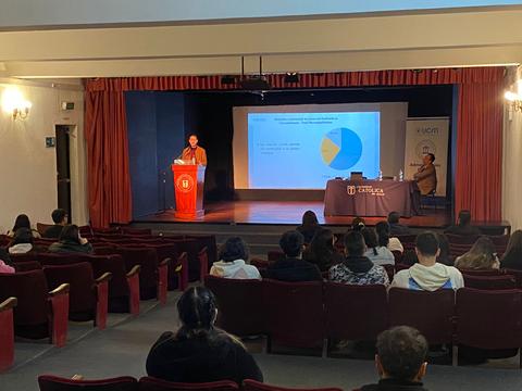 Una presentación en un auditorio con un ponente hablando frente a una pantalla y un público sentado.