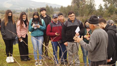Un grupo de jóvenes escucha atentamente a un hombre mayor que está dando una charla al aire libre sobre agricultura.