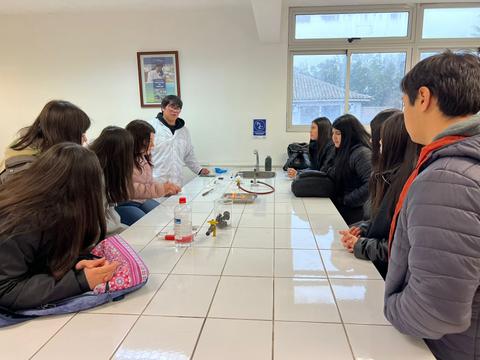 Un grupo de estudiantes se encuentra en un aula observando a un docente que les presenta un experimento en una mesa.