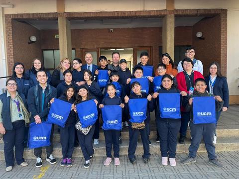 Un grupo de estudiantes y adultos posando juntos afuera de un edificio, todos sosteniendo bolsas azules con el logo de UCM.