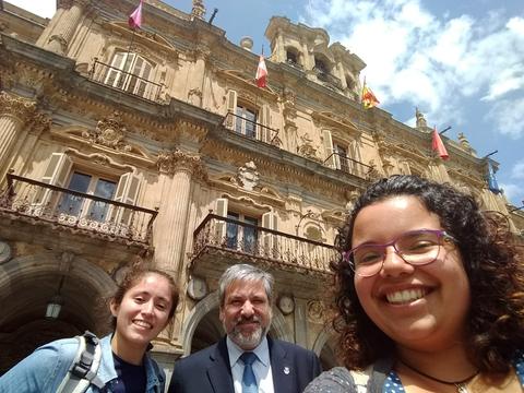 Tres personas sonríen frente a un edificio antiguo con banderas ondeando.