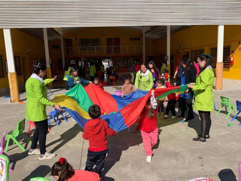 Un grupo de adultos y niños juega con una gran tela de colores en un patio escolar.